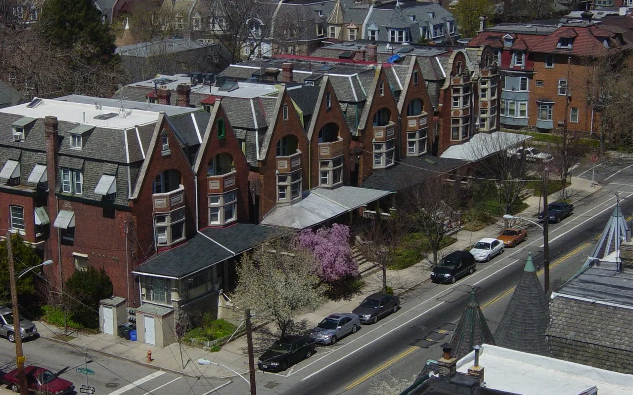 Brick rowhouses with porches and rooftop units along a tree-lined street in West Philadelphia
