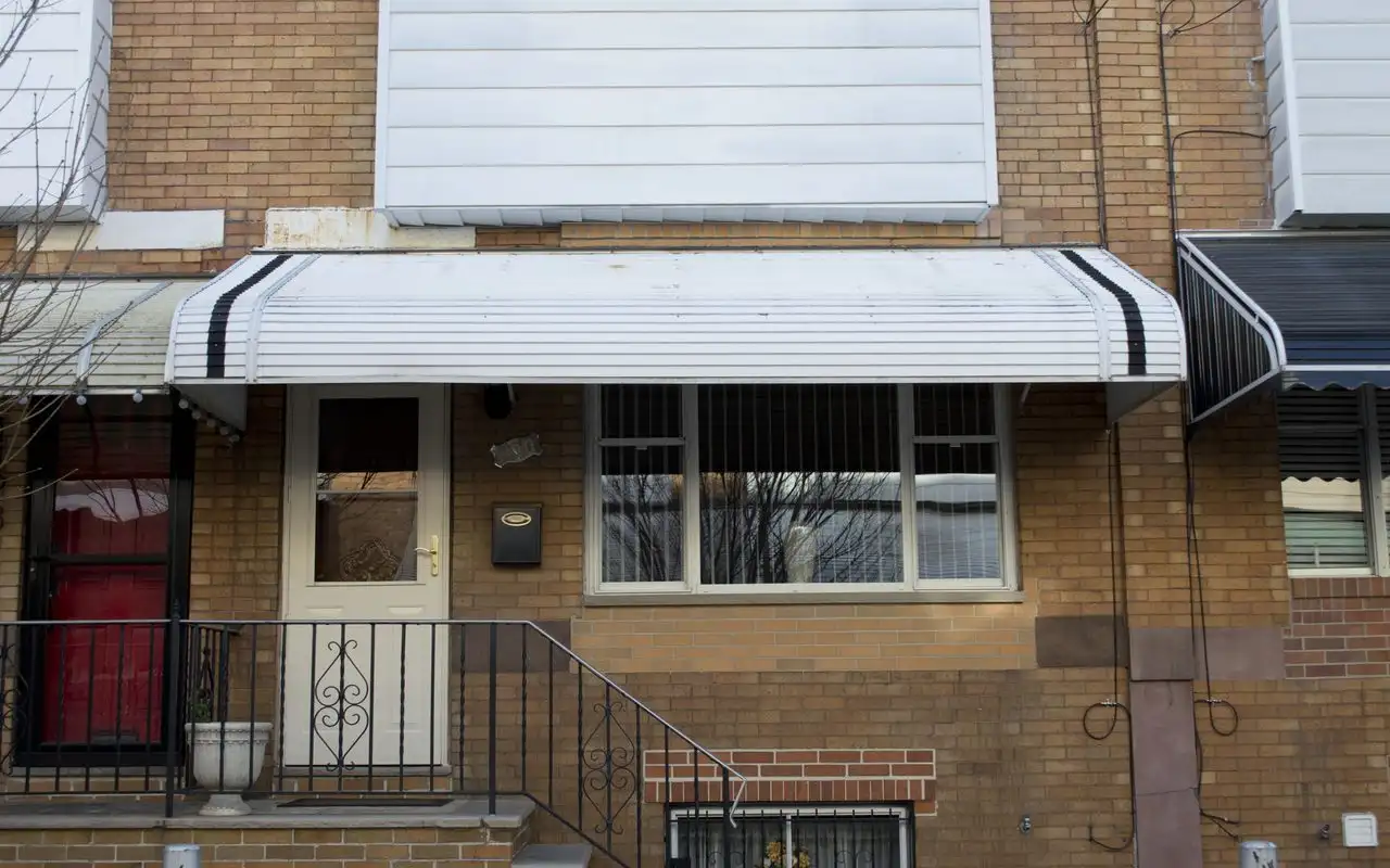The front of a brick South Philadelphia rowhouse with a white metal awning over the door and window and iron railings on the front steps